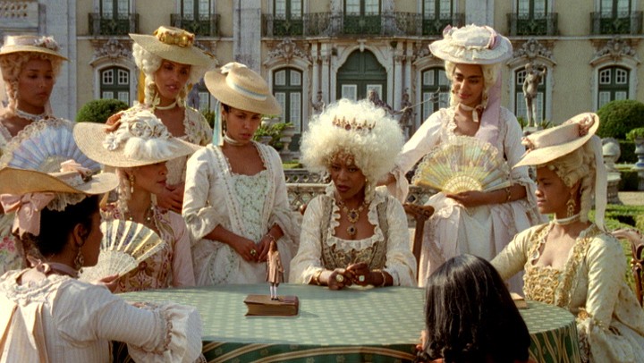 A still from Gullivers Travel, showing women sat at a table in period dress, with white wigs on