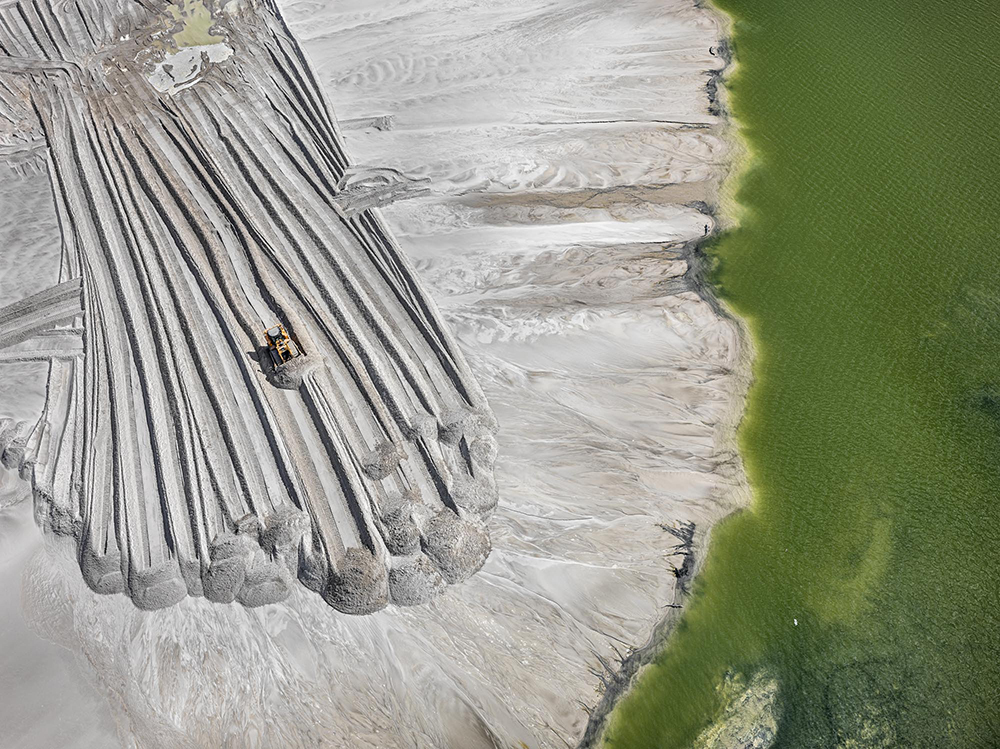 An aerial photo taken by Edward Burtynsky, Phosphor Tailings Pond #4, Near Lakeland, Florida, USA. On a white plane a digger pushes sediment or dirt to a pile. To the right is a green body of water. 