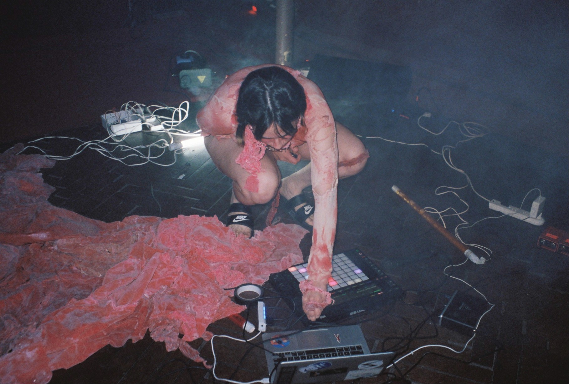 A photo of Candie performing. She is knelt on the floor making adjustments to her electronic instrument on the floor. There are swathes of pink fabric on the floor.