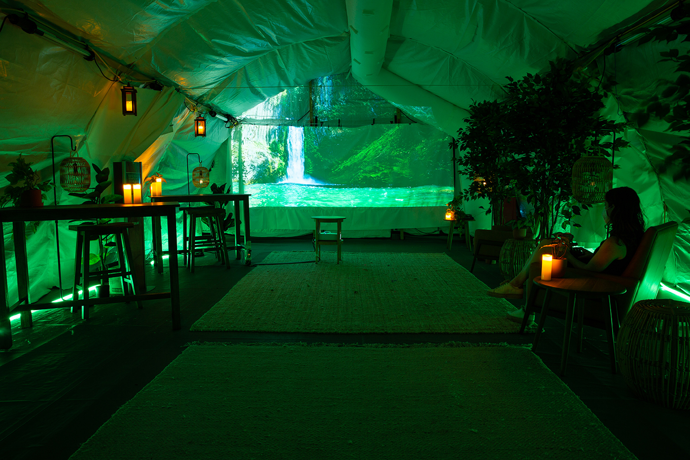 A photo representing New York. A cosy space washed in green, with someone curled up in an armchair to the right. The space is in a tent with candles, and at the end you can see a projection of a waterfall and greenery.