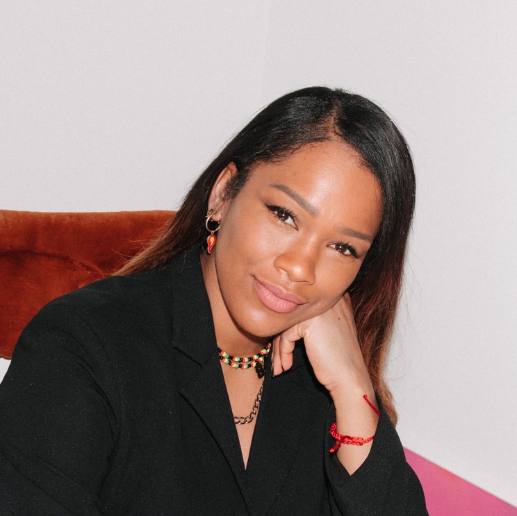 Headshot of Sharmadean Reid. Sharmadean is a Black woman with glossy straight hair, wearing a black blazer. She is leaning her cheek on her left hand and is smiling to the camera.