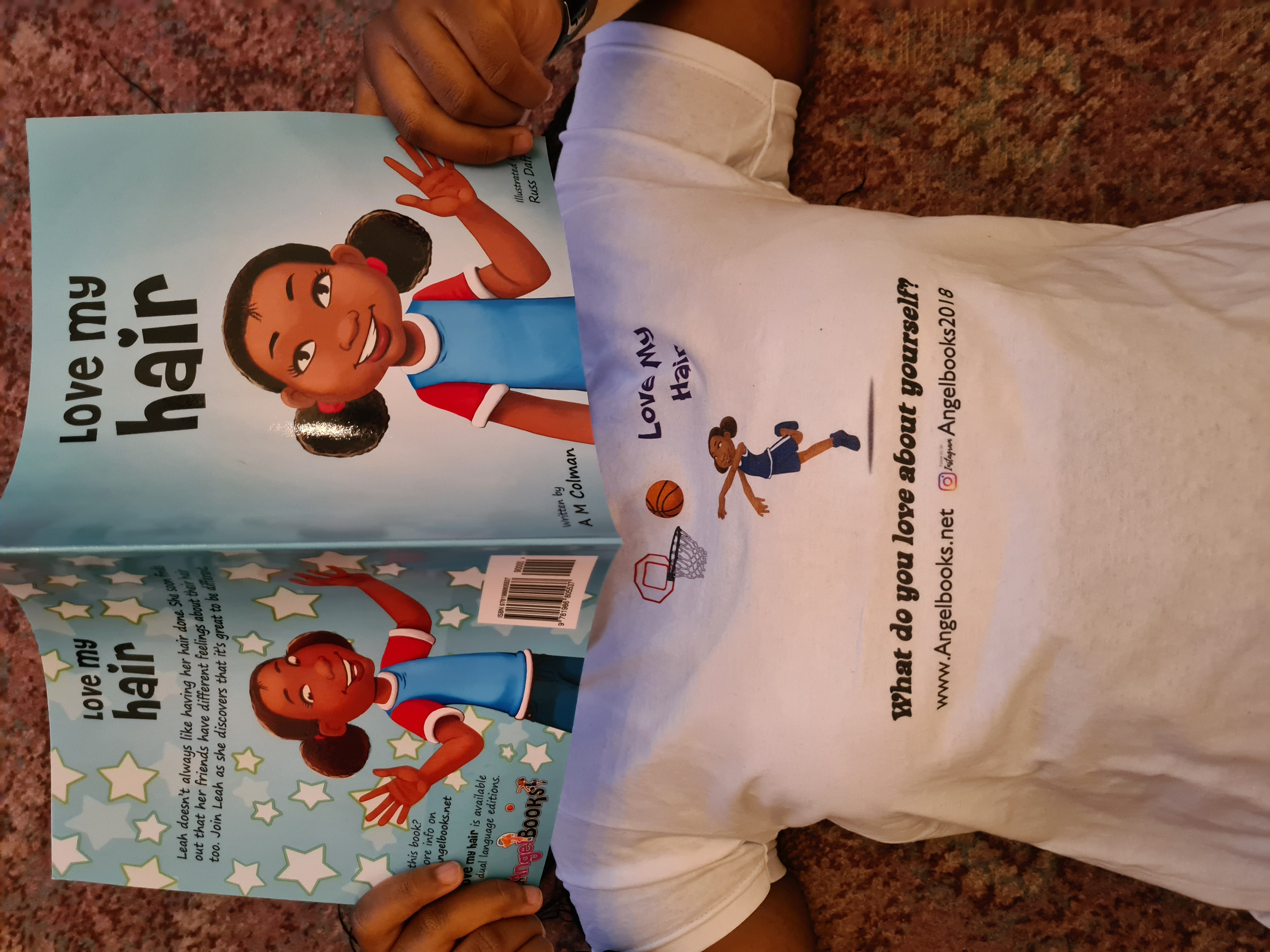 A Black girl holds a book in front of her. On the cover of the book ia a cartoon of a Black girl with the words 'Love my hair' written above.