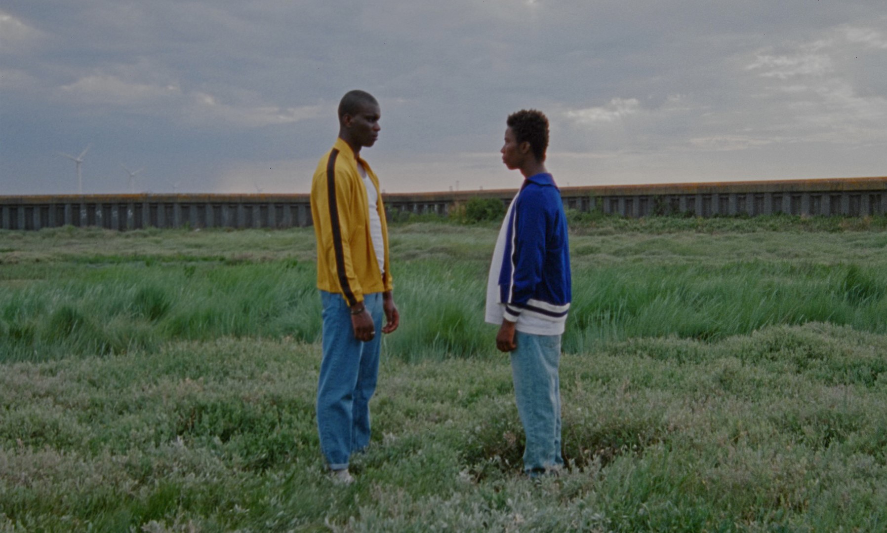 A still from a film by Iggy London. It shows two Black men, stood in a field, staring at each other's feet.