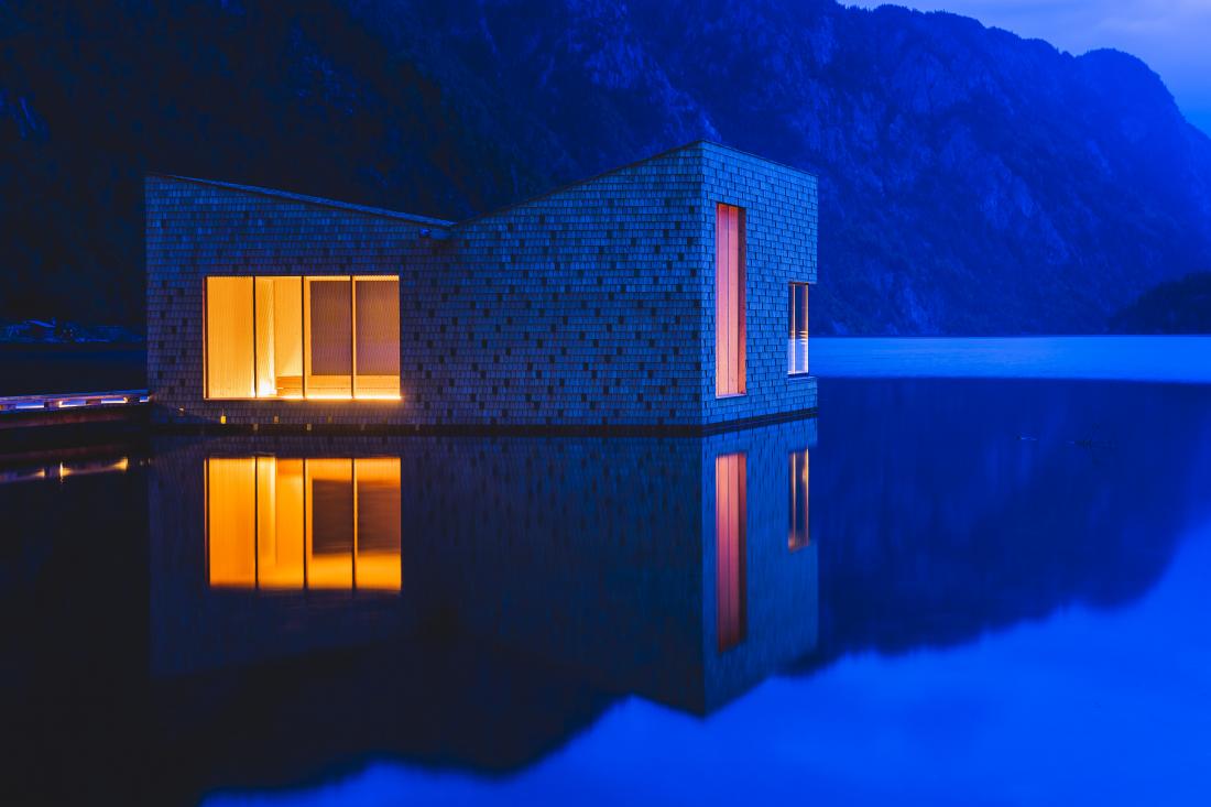 A photo of a minimalist house built on a calm body of water, with mountains in the background. The warm yellow lights coming from the window of the building contrast the calm blue of the water and mountains.