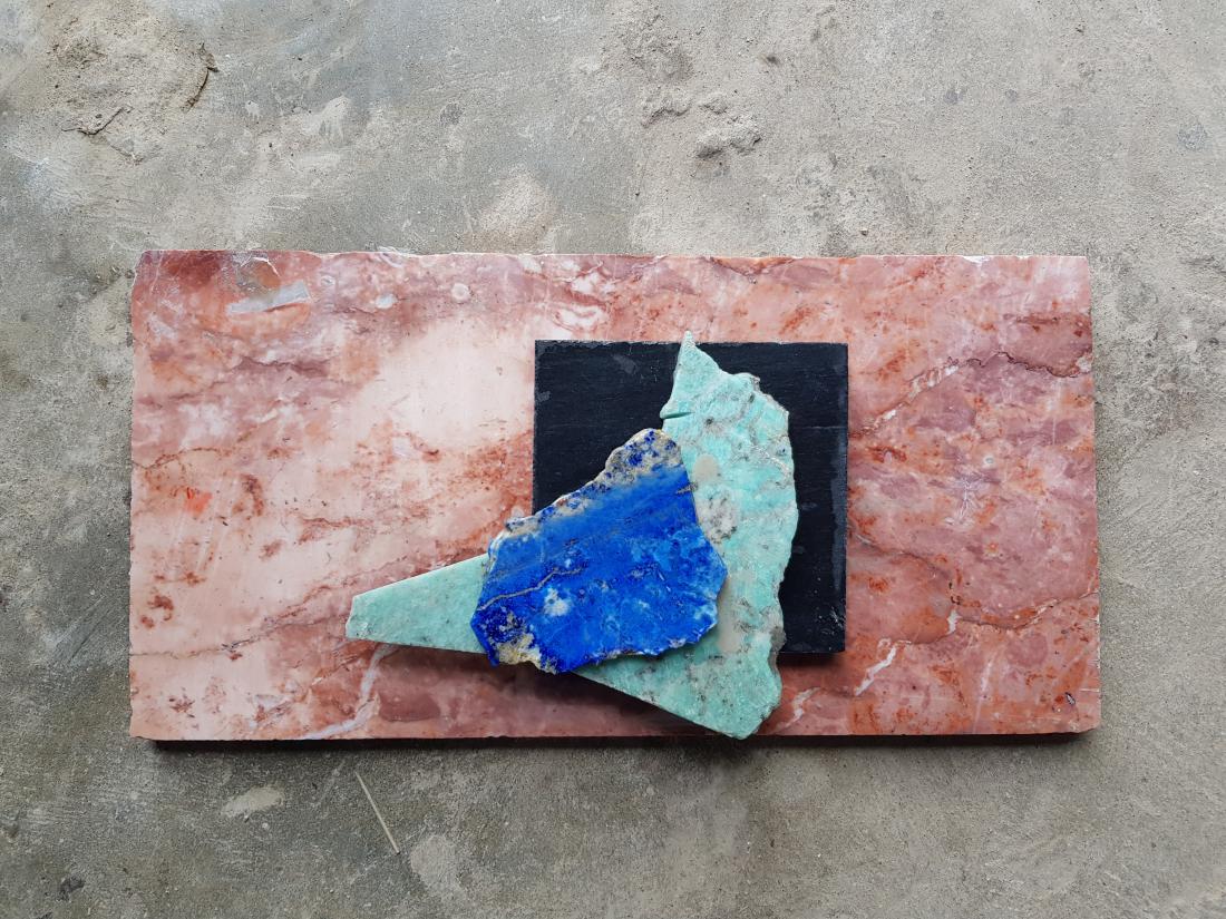 A photo representing Pakistan's Pavilion. There are layered textural samples - on the bottom is a grey stone. On top of this is a quartz like pink, which has a smaller navy blue shiny square on top. Layered on this is a triangular turquoise scrap and blue