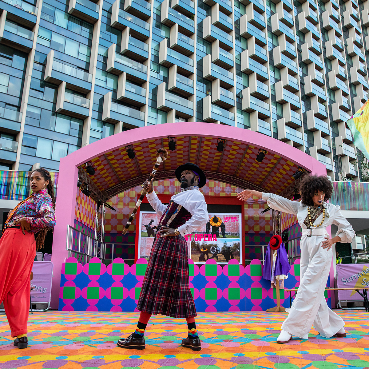 A photo of Patrick Ziza performing 'Dance by Design'. Four performers in Dandy clothing perform on stage. They are dressed in colourful shirts, hats and suit trousers, pulling suave poses.