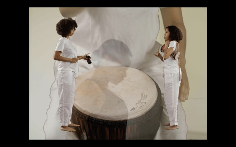 A still from Sonya Dyer's  video work 'Andromeda' (2021). Two women, wearing white, play hand-held percussion instruments. They face each other symmetrically . In the background you can see a faint outline of another woman, dancing, and a drum.