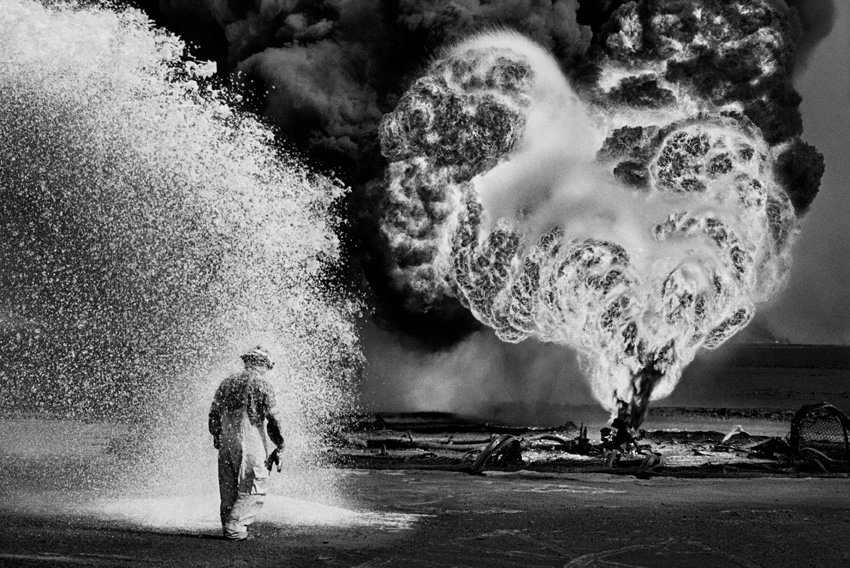A black and white image of a figure wrapped in protective fire fighting gear. They walk through a cloud of white liquid, while in the distance a well of oil burns fiercely, with great plumes of black smoke mushrooming into the sky,