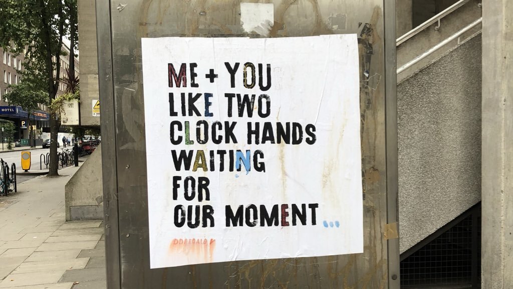 A photo of a paste-up poster on the side of a phone box. It reads Me + You like two clock hands waiting for our moment 