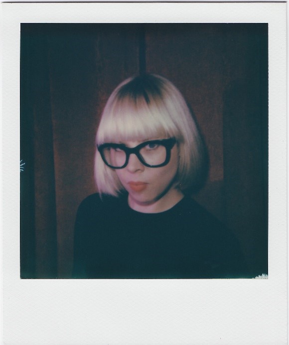 A polaroid of Nova Dando. She smiles and is looking to camera, with a blonde bob and fringe, with black, square glasses.