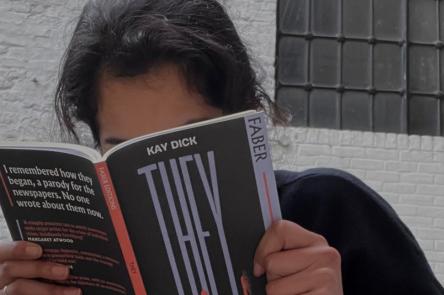 A photo of Aiysha reading a book called They by Kay Dick. The book has a black cover with a red spine, and obscured Aiysha's face.