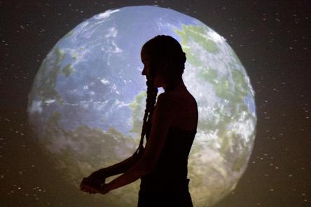 A woman (Anna Mikkola) standings in front of a projection of the world, as seen from space