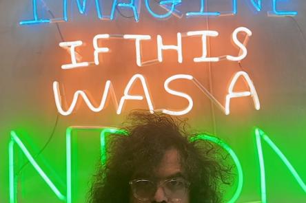 A photo of Babak Ganjei stood in front of one of his artworks. Babak stands at the front of the shot, and only his eyes and curly dark hair can be seen peering above. In the background is a neon artwork, which says 'Imagine if this were a neon'