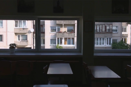 A still from Gerda Paliušytė's film Early Winter. It depicts a class room, with empty chairs and desks, with windows, looking out to buildings across the way.