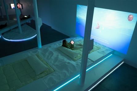An installation image of an artwork by the collective Keiken. In a blue-lit gallery space are projections and TVs on the wall. On the floor are pillowy mattresses and cushions, surrounded by sand. People are reclining on these structures.
