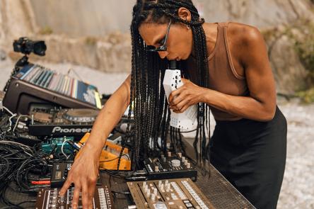 A photo of Nkisi performing on a beach at Nextones. Nkisi is shown in profile, with her decks and electronic sound equipment in front of her.