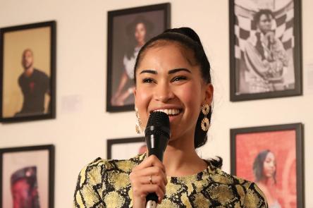 A photo of Swarzy smiling with a microphone in her hand. She stands in front of a wall of framed photos of Black creatives at an exhibition created by Swarzy, entitled Too Much Source.