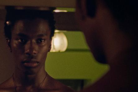 A still from a film by Iggy London. It shows a Black man, loking into a mirror, where we can see a reflection of his face.