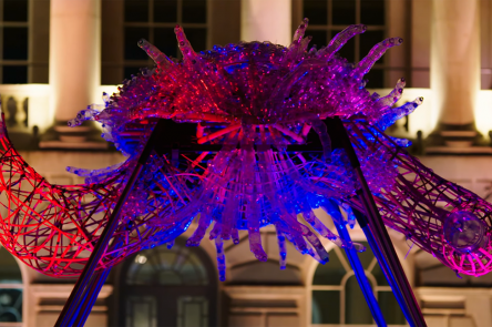 Close up of Leeroy New's installation The Arks of Gimokudan in the courtyard at Somerset House. It shows one of the sculptures, made up of reused plastic to look like a creature with arms, lit up by ambient lighting of pink, red and purple.