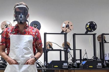 A photo of a man stood in front of 3-D printing equipment wearing a red shirt and a plastic face shield.