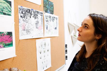 A photo of Mirelle Phillips looking at a pin board with focus. On the pin board are photos of trees alongside drawings and diagrams.