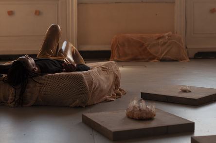 An installation of image 'We can no longer deny ourselves'. The artist lies on a fabric covered cushion with an arm above their head and knees in the air.  On the floor around them are low plinths with crystals neatly placed on them.