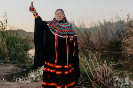 A woman wearing a long black dress and holding up a smoke cleansing bundle aloft in front of a body of water