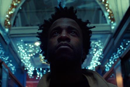 A still from the film Simeon Barclay: Defying the Norm. In it the artist Simeon Barclay, a Black man with medium length hair looks upwards as he stands in a shopping arcade in Leeds, that is lit with fairy lights in the darkness.