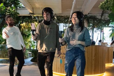 A photo of placement participants at work on 'Green Planet'. Two young people hold technical film equipment out in front of them, while someone else looks on with their arms thrown wide. There are green plants all around them.