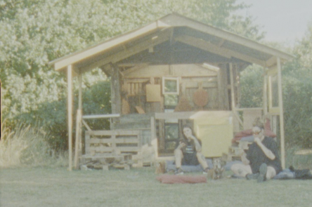 A still from Babeworld's 'Derby Day', 2022. Two people sit on the grass outside a wood cabin. The scene is shot on fuzzy, atmospheric film. 