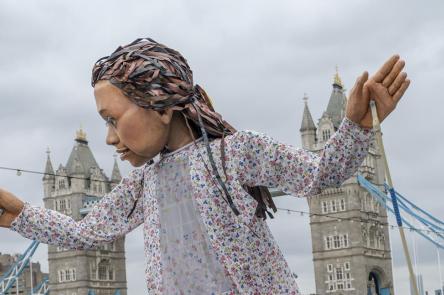 A photo of Little Amal, walking alongside the River Thames. In the background you can see Tower Bridge. The puppet looks to its right, with her hands held up and apart.