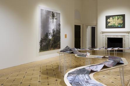 A photo of an installation view of We Are History  In the middle of the room is a work laid out on a twisting table, like the bends of a river.  On the wall are two artworks depicting tropical scenes, and an orange sign that says We Are History.