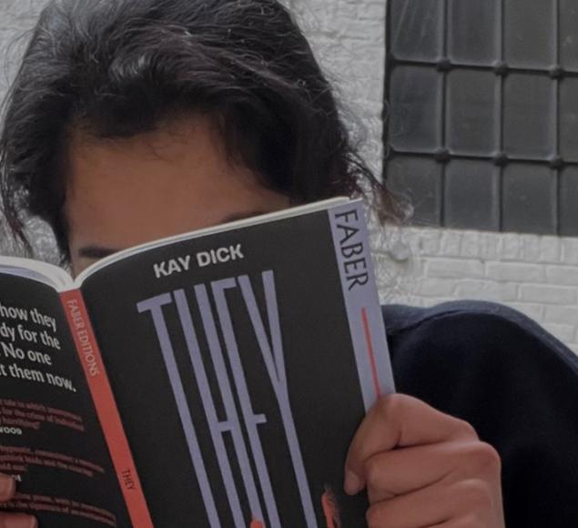 A photo of Aiysha reading a book called They by Kay Dick. The book has a black cover with a red spine, and obscured Aiysha's face.