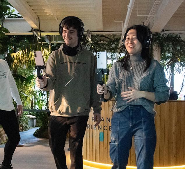 A photo of placement participants at work on 'Green Planet'. Two young people hold technical film equipment out in front of them, while someone else looks on with their arms thrown wide. There are green plants all around them.