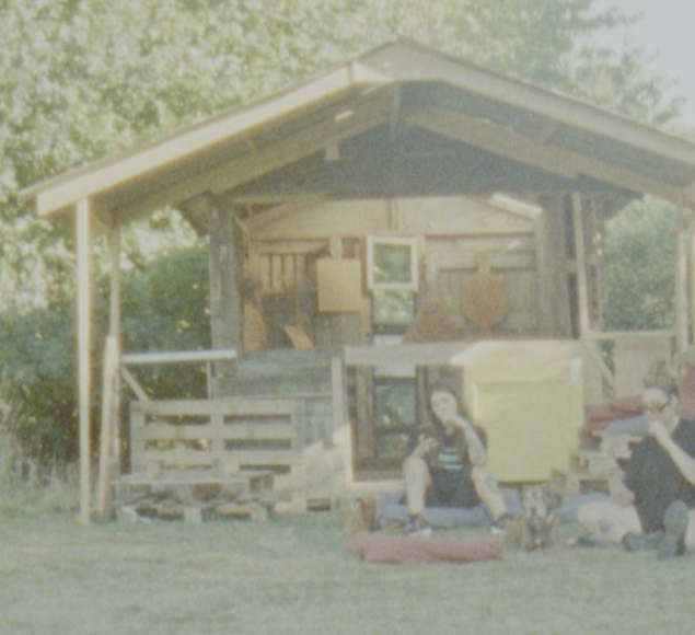 A still from Babeworld's 'Derby Day', 2022. Two people sit on the grass outside a wood cabin. The scene is shot on fuzzy, atmospheric film. 