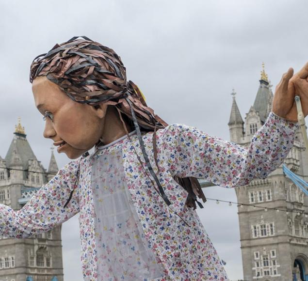 A photo of Little Amal, walking alongside the River Thames. In the background you can see Tower Bridge. The puppet looks to its right, with her hands held up and apart.