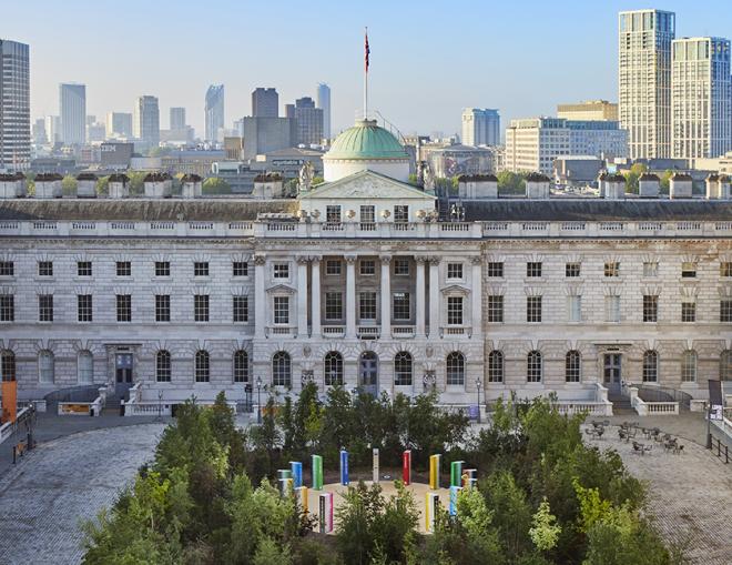 Forest for Change at Somerset House. Green leafy trees fill the courtyard. In the background you can see the London skyline and blue skies.