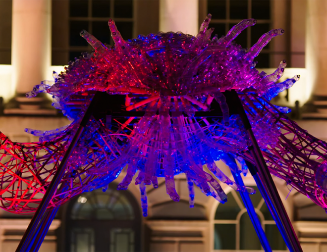 A photo of Leeroy New's installation The Arks of Gimokudan in the courtyard at Somerset House. It shows one of the sculptures, made up of reused plastic to look like a creature with arms, lit up by ambient lighting of pink, red and purple.