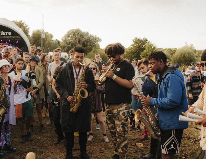A photo of Tomorrow's Warriors performing at We Out Here Festival. A group of musicians perform on brass instruments while a crowd gathers arou.d