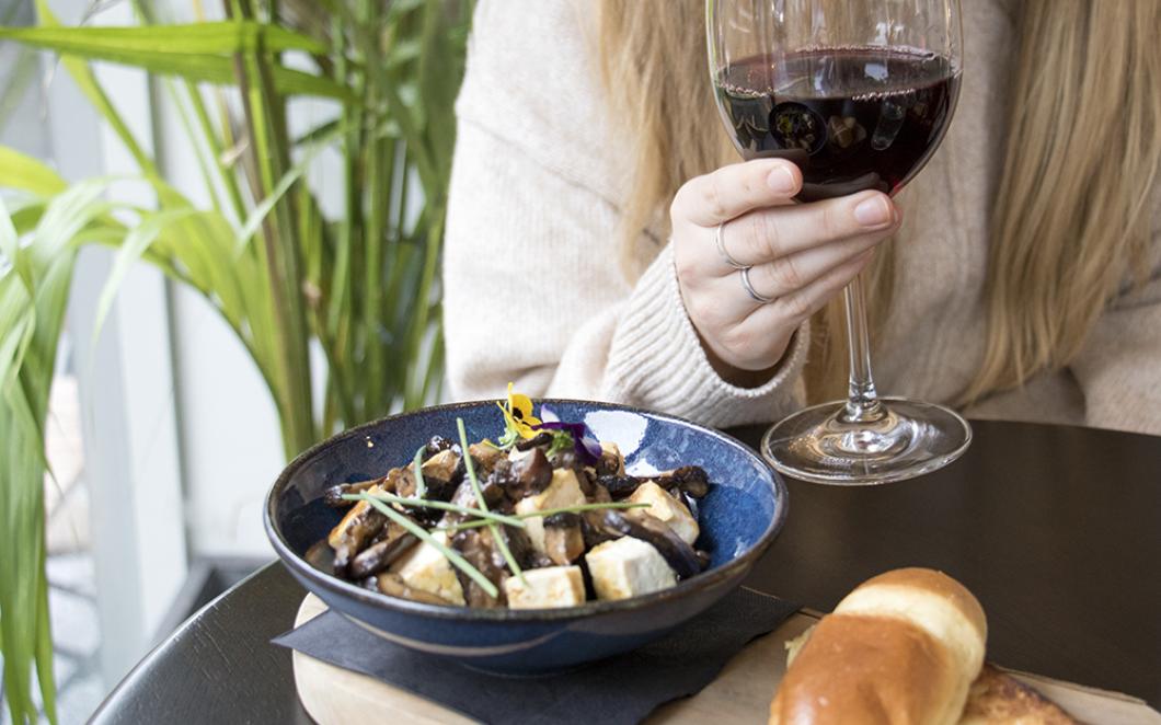 A bowl of shiitake mushrooms with soya glaze and smoked tofu on a table where a woman sits holding a glass of red wine at Grays & Feather