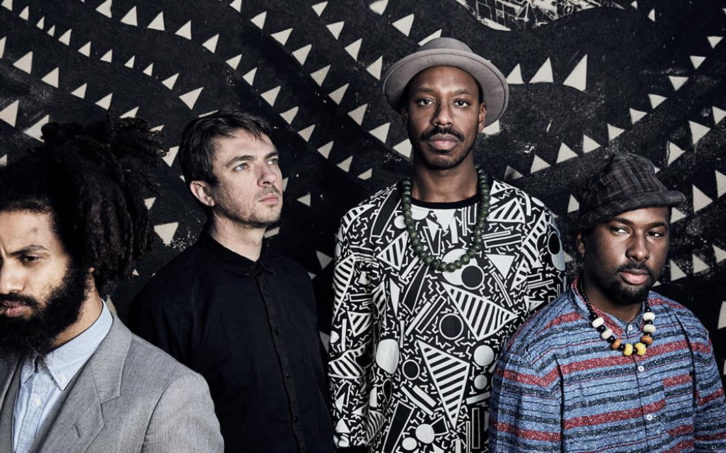 A photo of the band Sons of Kemet. Four men, stood against a patterned black and white backdrop.