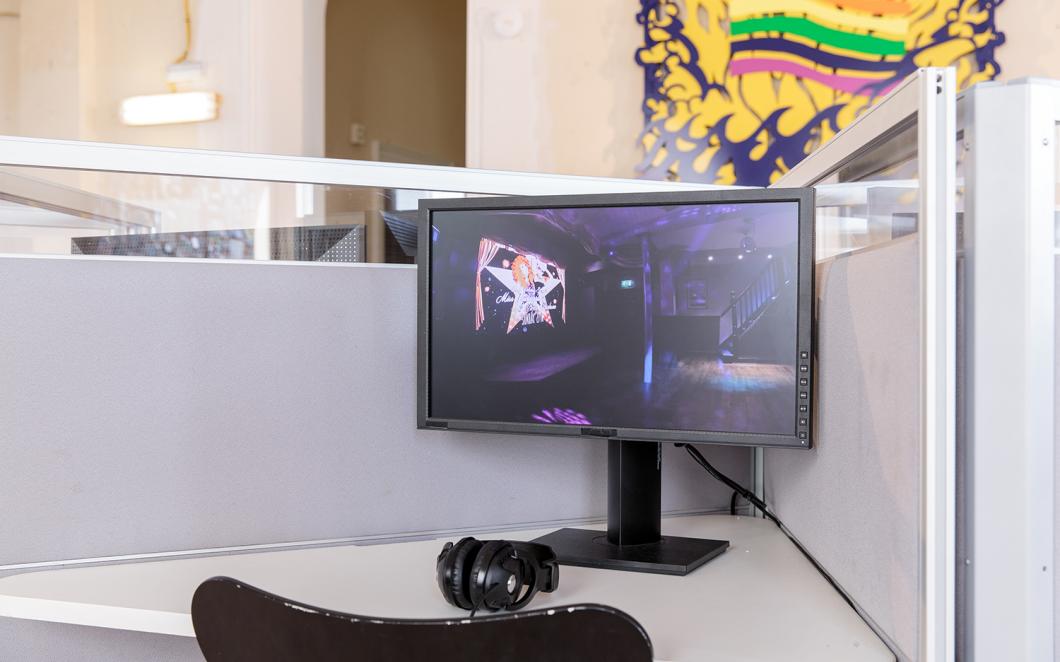 Installation view of UK Gay Bar Directory. The image shows a computer and screen at a desk. Behind there is an artwork on the wall showing a smiling sun and a rainbow flag.