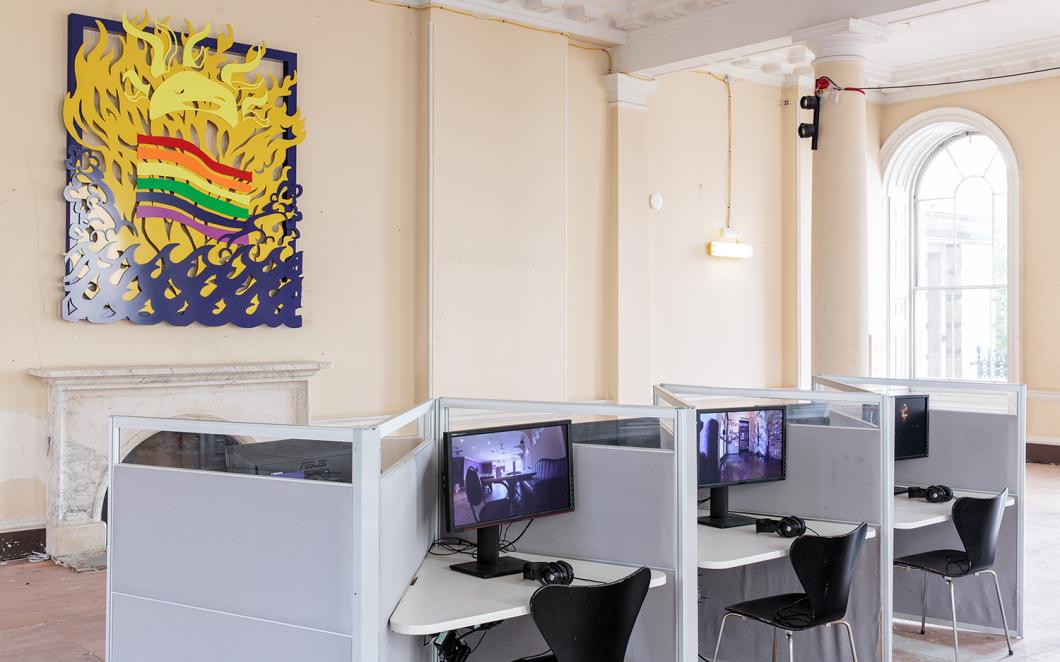 Installation view of UK Gay Bar Directory. The image shows 3 computers at desks. Behind there is an artwork on the wall showing a smiling sun and a rainbow flag.