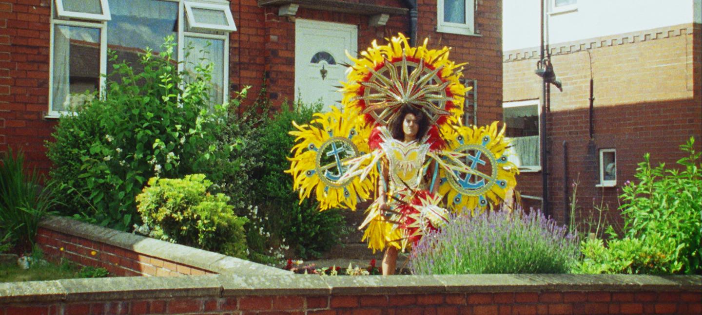 An image of a woman stood in a bright, colourful yellow, sun-like costume in a front garden.