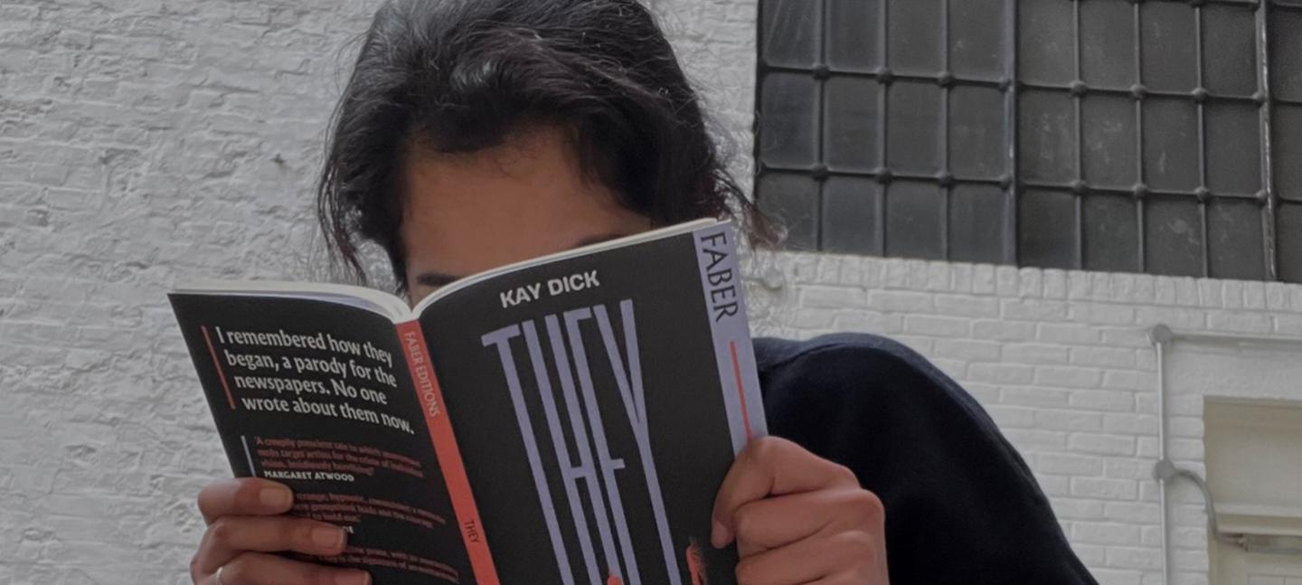 A photo of Aiysha reading a book called They by Kay Dick. The book has a black cover with a red spine, and obscured Aiysha's face.