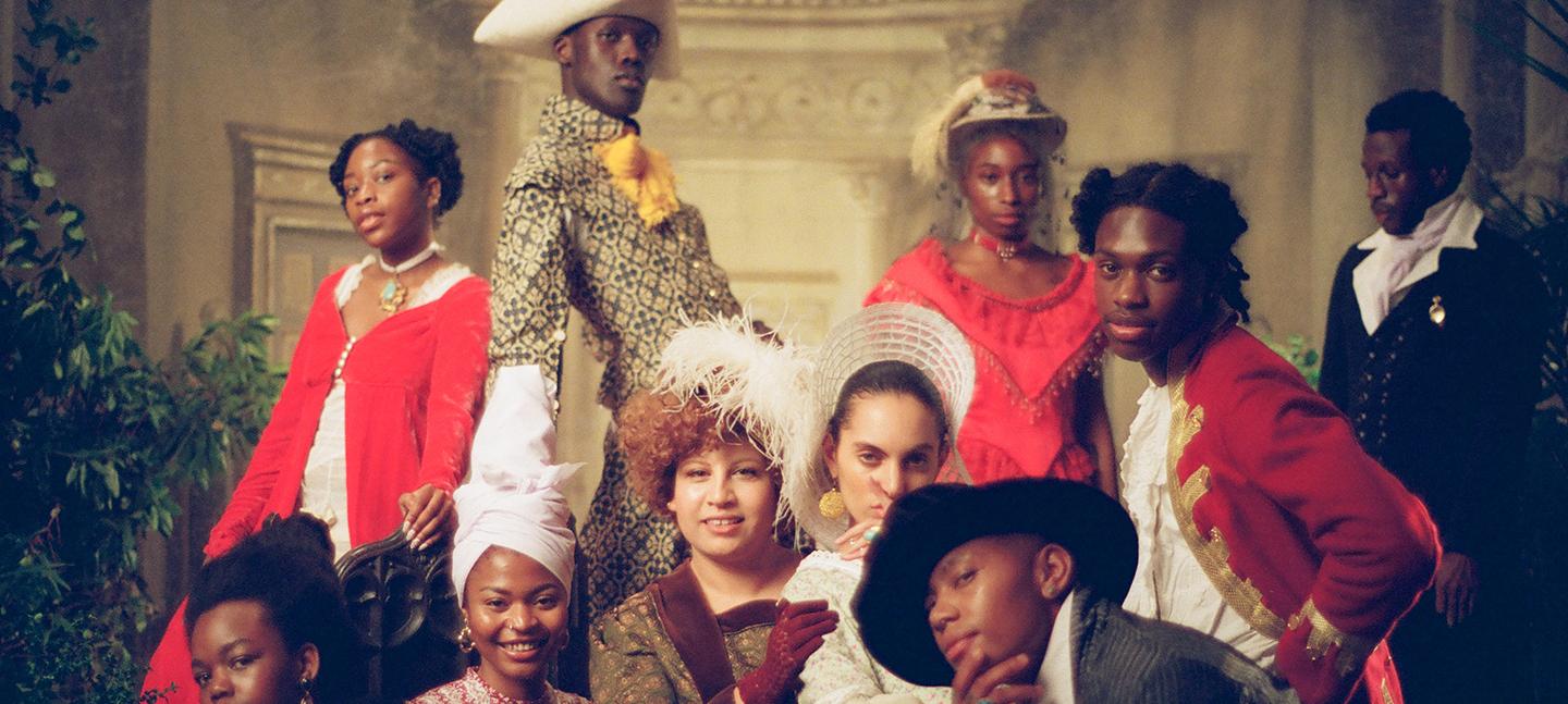 A still from an Akinola Davies Jr. film. It shows young people of colour dressed in Regency period dress, wearing suits with feathers in their hats and long sweeping gowns. They are all looking to camera and smiling.