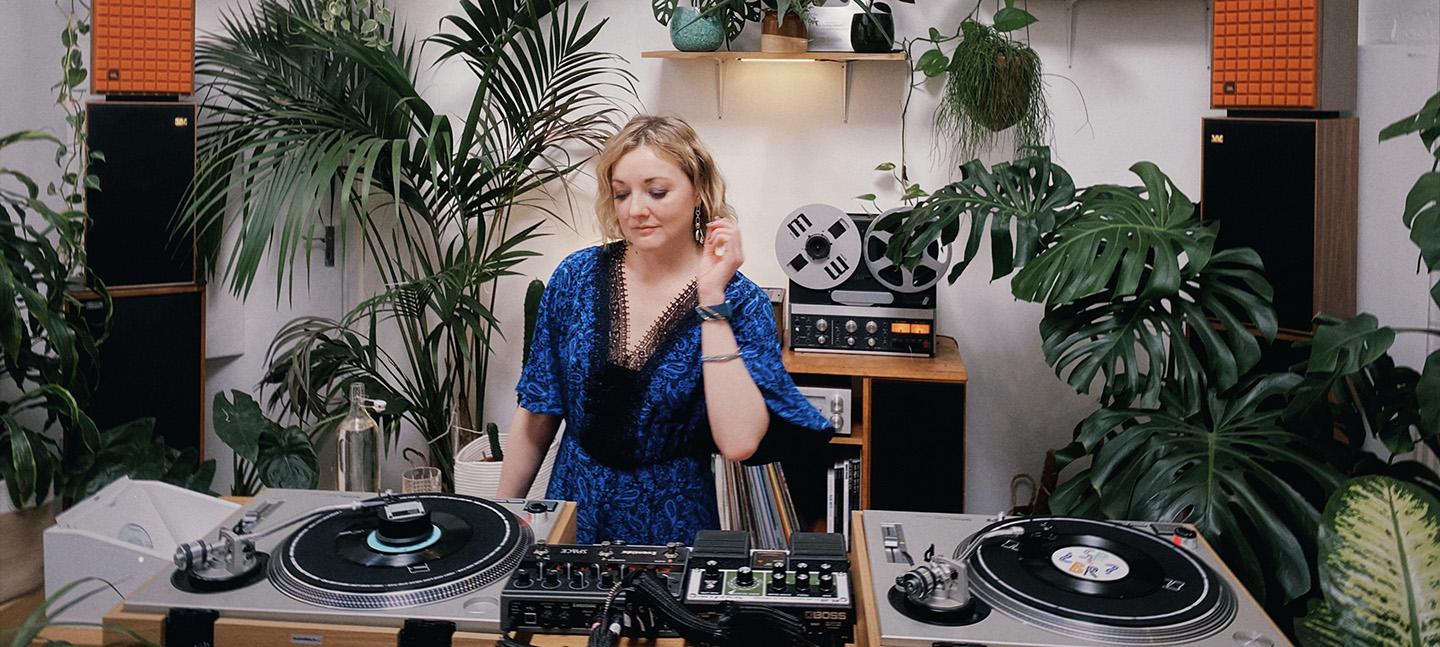 Alena Arples is at work on the decks. She is DJing in a light-filled room full of plants.