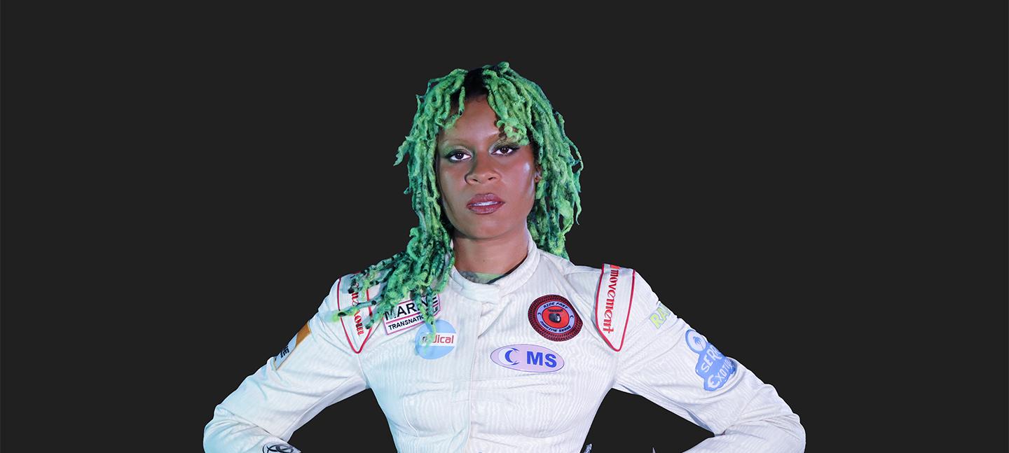 The musician Aluna is a Black woman who stands wearing a white one-piece suit with hands on her hips. Her hair is vibrant green braids and she looks directly to camera.