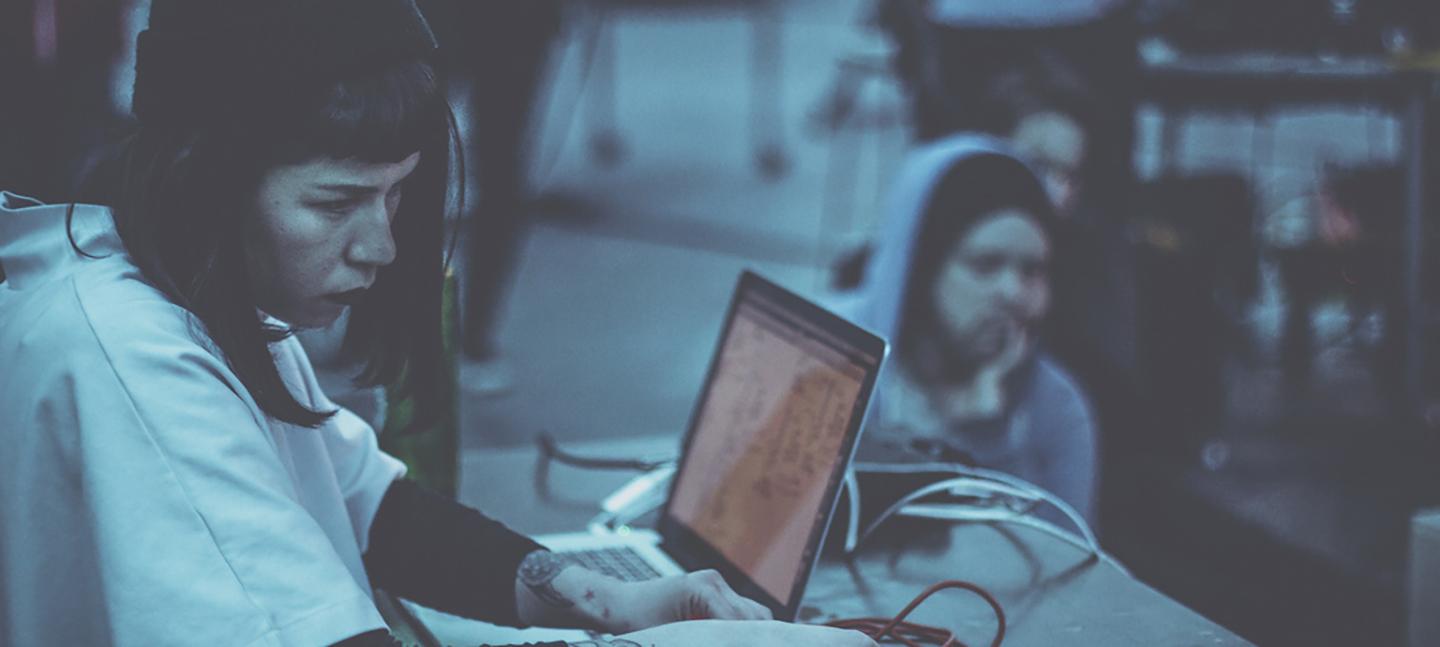 A photo of Chloe Alexandra McKenzie at her laptop, while people sit around her listening.