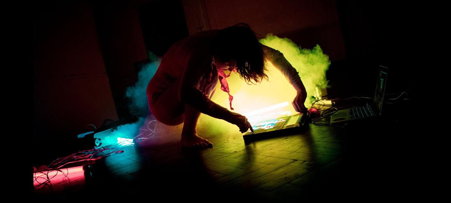 A photo of Candie performing. She is knelt on the floor making adjustments to her electronic instrument on the floor. There are pink, blue and yellow lights around her.
